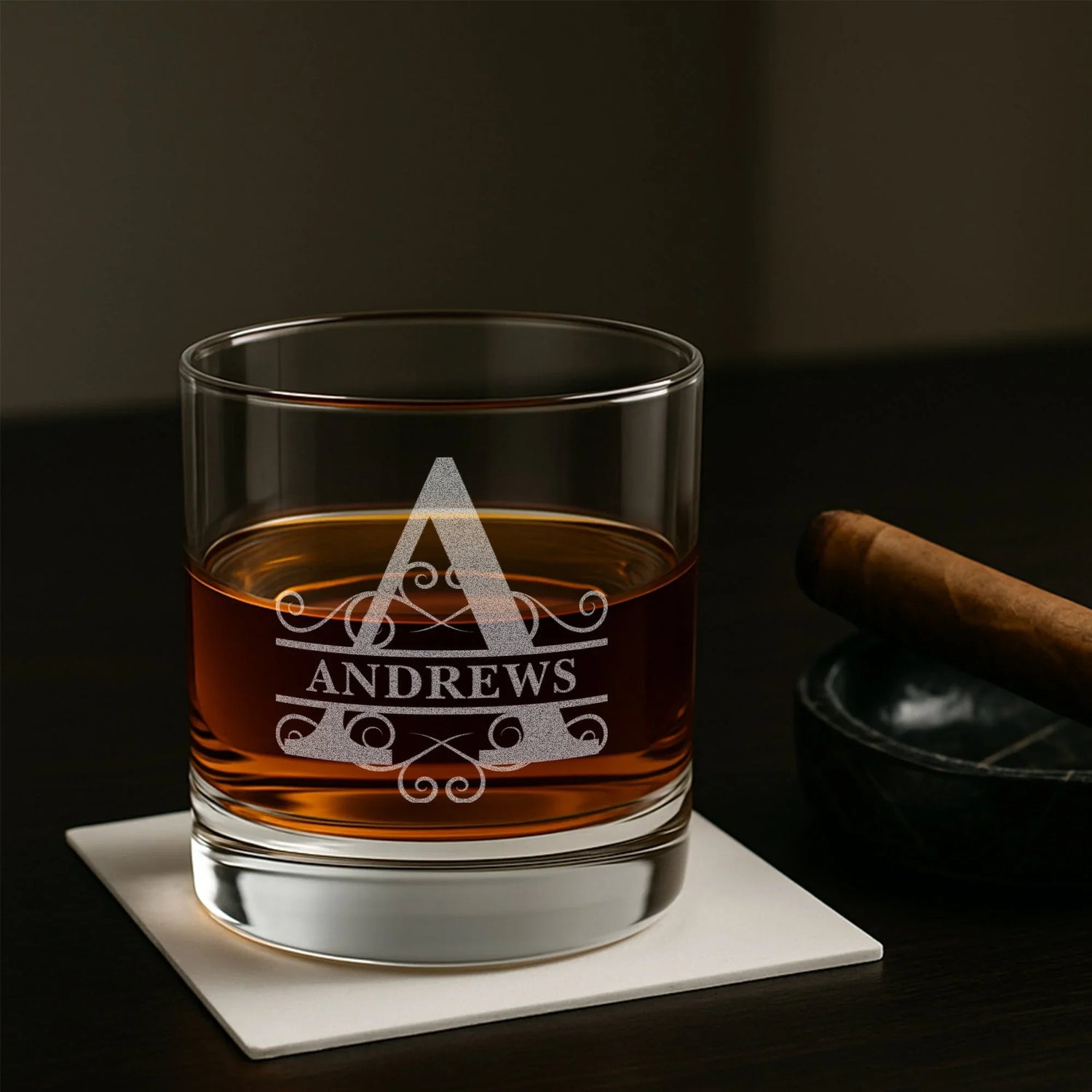 Personalized etched whiskey glass with the name Andrews, beside a cigar on a dark table.