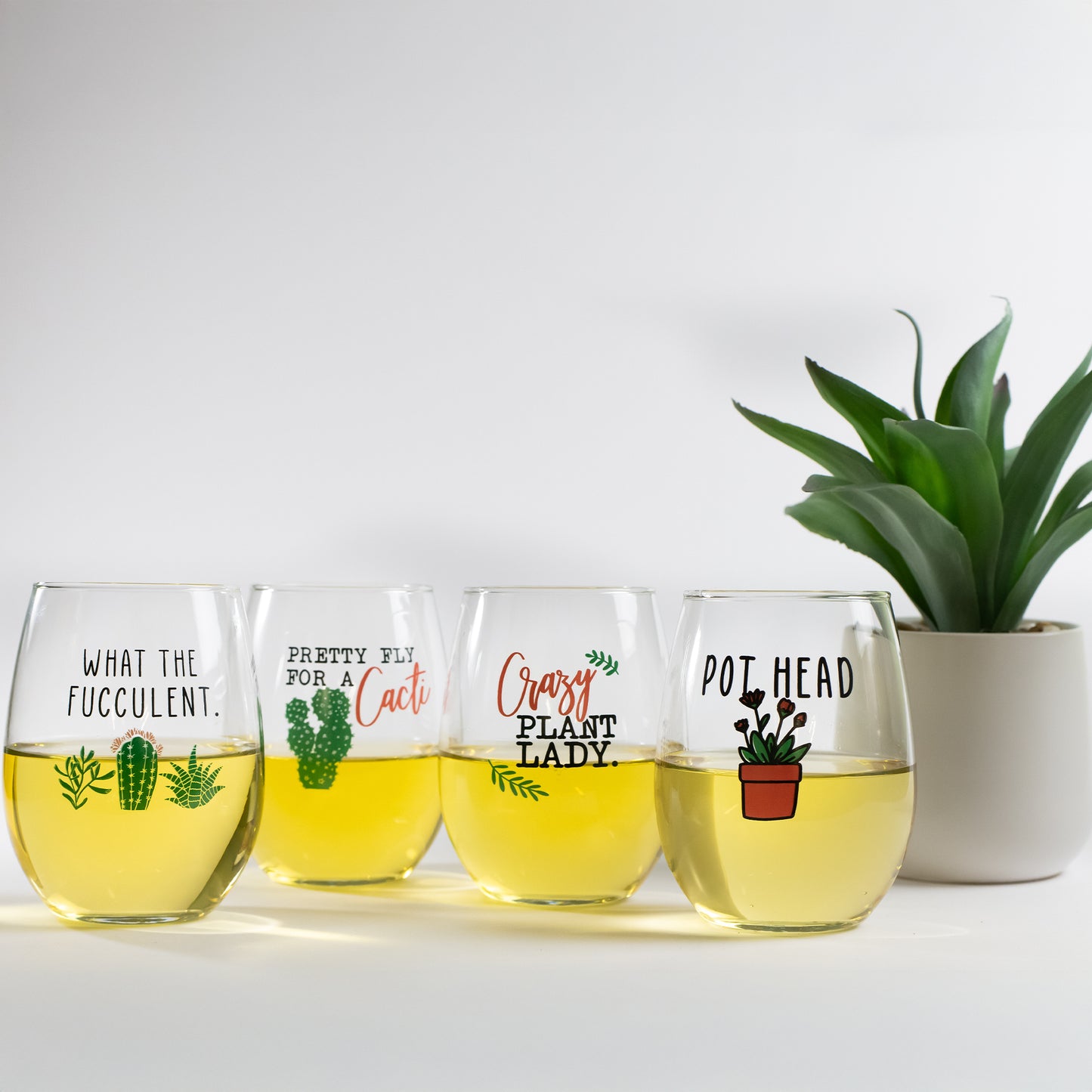 Set of four stemless wine glasses with funny succulent and cactus quotes, next to a potted plant.