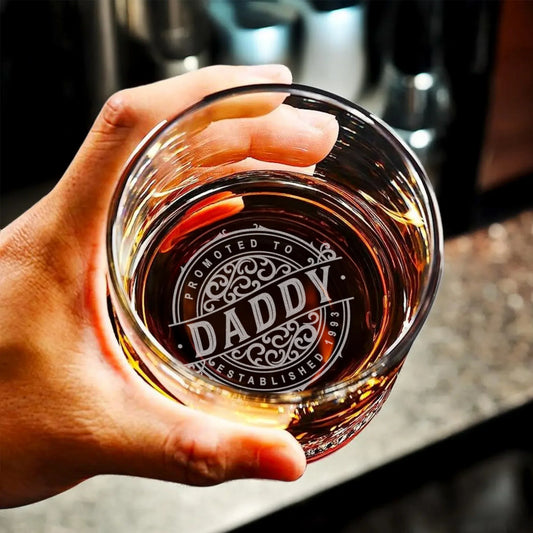 Hand holding personalized whiskey glass engraved with 'Promoted to Daddy' over dark drink.