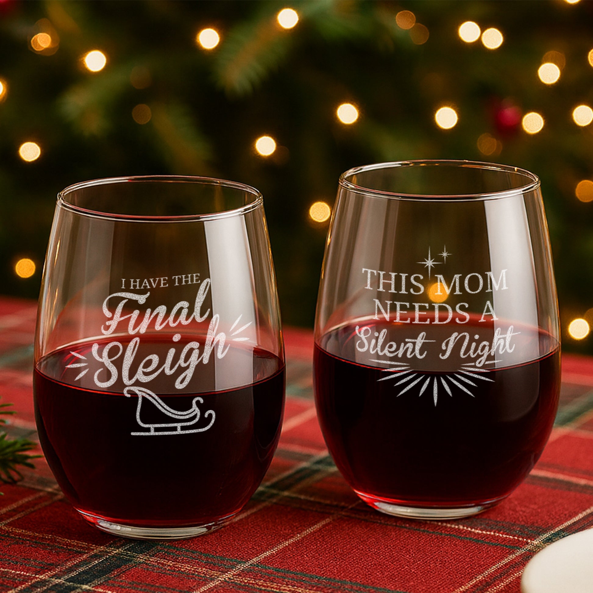 Two engraved Christmas wine glasses with festive sayings, filled with red wine on plaid tablecloth.